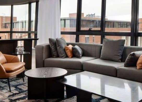 An open plan living room in grey & brown motif with huge windows looking out to buildings in the surrounding.