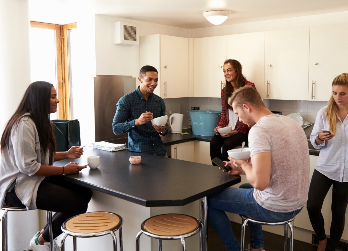 People standing and sitting around a kitchen table in a student accommodation.